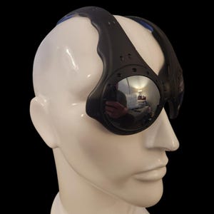 May include: Black, futuristic-style goggles with a reflective lens. The goggles have a curved, black frame that wraps around the head. The lenses are circular and reflect the surrounding environment. The goggles are on a white mannequin head.