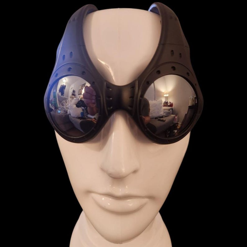 May include: Black steampunk-style goggles with round, reflective lenses. The goggles have a thick, curved frame with rivet details and a bridge connecting the lenses. The goggles are displayed on a white mannequin head against a black background.