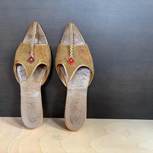 May include: A pair of gold-colored, open-toe sandals with a decorative pattern. Each sandal features a red gem and a braided strap. The sandals are displayed on a light wooden surface against a dark background.