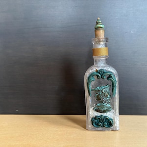 May include: A clear glass bottle with a decorative teal and bronze design. The bottle has a cork stopper and a gold band around the neck. The design features a profile of a person in a hat.