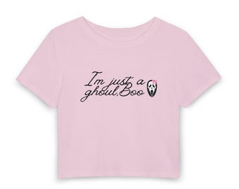 May include: A light pink short-sleeved crop top featuring the black text "I'm just a ghoul, Boo" printed on the center chest. The design includes a small graphic of a white ghost mask with a tiny pink bow positioned above the word "Boo".