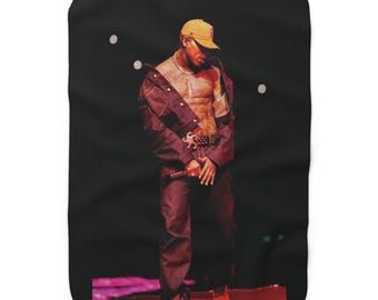 May include: A rectangular fleece blanket featuring a printed image of a person wearing a mustard yellow baseball cap, an unbuttoned dark denim jacket, and matching dark denim pants. The person is shirtless, revealing a tattoo on the chest.