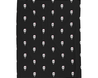 May include: A black blanket featuring a repeating pattern of white ghost-like masks, each adorned with a small pink bow on the forehead. The design is evenly spaced across the entire surface of the fabric, creating a consistent graphic print.