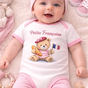 May include: A smiling baby wearing a white onesie with pink trim and the words "Petite Française". The onesie features a cartoon bear with a beret, holding a baguette and a French flag. The baby is also wearing a pink headband and pink pants.