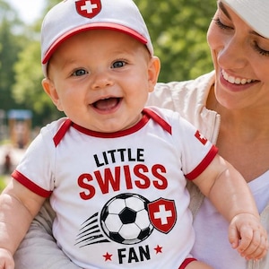 May include: A baby in a white onesie, cap, knee pads, and trainers, all featuring Swiss flag emblems. The onesie says "Little Swiss Fan" with a football graphic. The baby is held by a person wearing a matching cap.