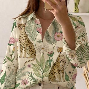 May include: Cream-colored button-down shirt with a vibrant floral and cheetah print. The long-sleeved shirt features pink flowers, green leaves, and two cheetahs. The shirt is paired with white jeans.