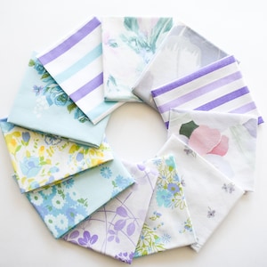 May include: An overhead shot displays a collection of folded vintage fabric squares arranged in a circular pattern. The fabrics feature floral and striped patterns in pastel colors like blue, purple, and yellow. The arrangement is on a white surface.
