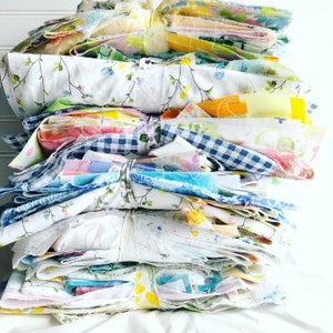 Pode incluir: Uma pilha de pacotes de tecido amarrados com barbante. Os tecidos apresentam padrões florais, xadrez e lisos em tons pastel. Os pacotes parecem ser feitos de têxteis vintage reaproveitados, perfeitos para projetos de artesanato ou colchas.