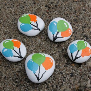 May include: Five white painted rocks with colorful balloon designs. Each rock features a cluster of three balloons in blue, green, and orange, connected by black stems. The rocks are arranged on a textured, gray surface.