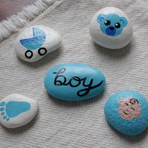 May include: Five painted rocks with baby-themed designs. One rock has a blue baby carriage, another has a blue footprint, and a third has the word "boy" in cursive. The other two rocks have a baby face and a bear face.