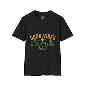 Può includere: T-shirt nera con la scritta "Good Vibes & Bad Throws" in oro, verde e viola. Il design include grafiche di gigli e maschere, perfetto per le celebrazioni del Mardi Gras.