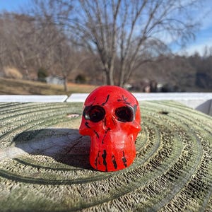 May include: A small, red skull figurine with black accents. The skull has dark eye sockets and is positioned on a weathered, circular wooden surface. The background features a blurred view of trees and a bright sky.