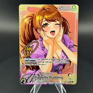 May include: A Charlotte Pudding trading card from the One Piece card game featuring a character with brown hair in pigtails, wearing a purple top, with a gold signature overlay. The card displays 5000 power, 4 life, and game text.