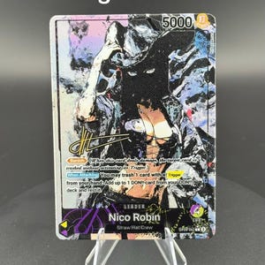 May include: A One Piece trading card featuring Nico Robin as a leader with 5000 power and 4 life. The card displays a stylized illustration of the character in a hat, a gold signature, and text detailing Banish and When Attacking abilities.