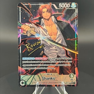 May include: A One Piece trading card featuring the character Shanks with red hair, wearing an open white shirt and a black coat, holding a sword. The card displays the text 'Shanks', 'Leader', '5000', '5 Life', and 'Rovrov' in a signature style.