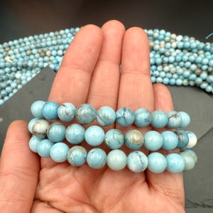 Puede incluir: Un hilo de cuentas redondas y pulidas de color azul claro con vetas jaspeadas en gris y blanco. Las cuentas est&aacute;n ensartadas en una l&iacute;nea continua, mostrando una textura suave y brillante y una forma esf&eacute;rica constante en toda la longitud.