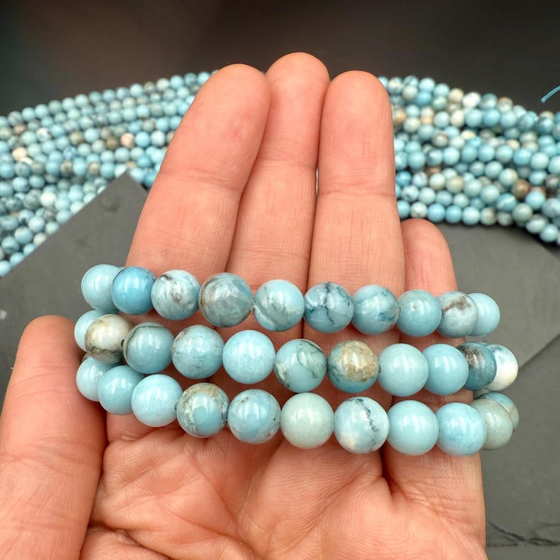 Puede incluir: Un hilo de cuentas redondas y pulidas de color azul claro con vetas jaspeadas en gris y blanco. Las cuentas est&aacute;n ensartadas en una l&iacute;nea continua, mostrando una textura suave y brillante y una forma esf&eacute;rica constante en toda la longitud.
