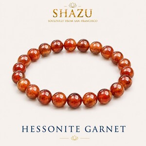 Pode incluir: Uma pulseira circular composta por contas de granada hessonita redondas, polidas e transl&uacute;cidas de cor laranja acastanhada. O texto SHAZU SOULFULLY FROM SAN FRANCISCO aparece no topo e HESSONITE GARNET est&aacute; impresso na base.