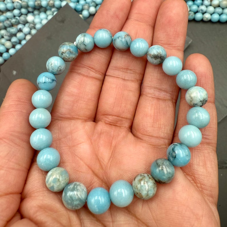 Puede incluir: Una pulsera el&aacute;stica compuesta por cuentas redondas y pulidas en varios tonos de azul claro y turquesa. Cada cuenta presenta patrones de veteado natural &uacute;nicos con sutiles inclusiones grises y blancas, creando un acabado suave y brillante.