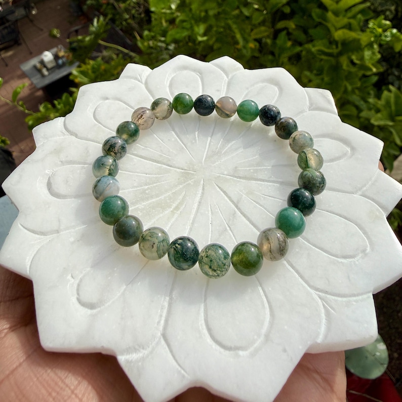 May include: A stretch bracelet composed of round moss agate beads featuring various shades of green, translucent white, and dark inclusions. The beads are polished to a smooth finish and arranged in a circular pattern on a white lotus-shaped base.