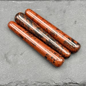 May include: Three polished obsidian wands with a reddish-brown base color and black speckles. The wands are cylindrical with rounded ends, lying diagonally on a dark gray surface. The wands are smooth and reflect light.