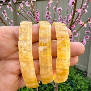 Puede incluir: Tres pulseras de piedras preciosas de color amarillo-naranja. Cada pulsera está compuesta por piedras rectangulares y facetadas. Las pulseras se sostienen en una mano, con un fondo borroso de flores rosas y follaje verde.