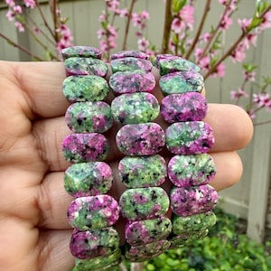 Puede incluir: Una pila de pulseras de piedras preciosas con cuentas ovaladas. Las cuentas son una mezcla de colores verde y magenta, con un patrón moteado. Las pulseras se sostienen sobre un fondo de follaje verde y flores rosas.