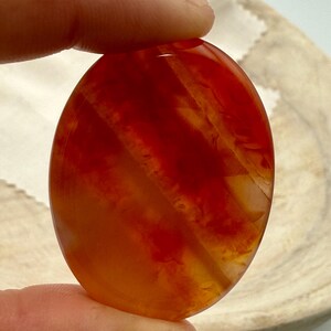 May include: A polished, oval-shaped gemstone with a vibrant blend of red and orange hues. The stone features natural patterns and striations, creating a unique visual texture. The gemstone is held between two fingers.