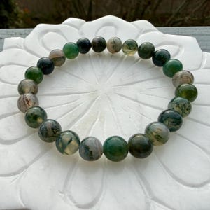 May include: A circular beaded bracelet featuring polished, spherical moss agate stones. The beads display a variety of translucent and opaque shades, ranging from deep forest green to earthy brown and creamy white, with natural moss-like inclusions.