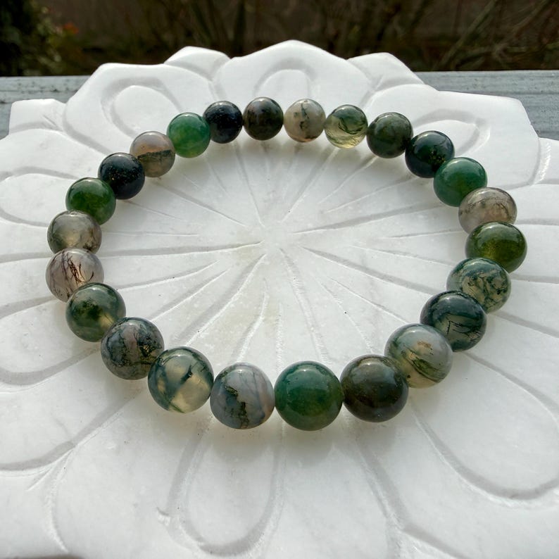 May include: A circular beaded bracelet featuring polished, spherical moss agate stones. The beads display a variety of translucent and opaque shades, ranging from deep forest green to earthy brown and creamy white, with natural moss-like inclusions.