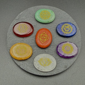 May include: Seven oval-shaped chakra stones in various colors, including red, orange, yellow, green, blue, purple, and white. Each stone features a gold-colored chakra symbol. The stones are arranged on a dark gray, circular surface.