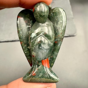 May include: A carved angel figurine made of green and red speckled stone. The angel has outstretched wings and a serene expression. The stone has a polished finish, and the figurine is held by a person's fingers.