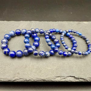 May include: Four stretch bracelets made of round lapis lazuli beads in varying sizes. The beads feature deep blue tones with subtle flecks of gold pyrite and white calcite inclusions, arranged in a circular pattern on a textured gray surface.