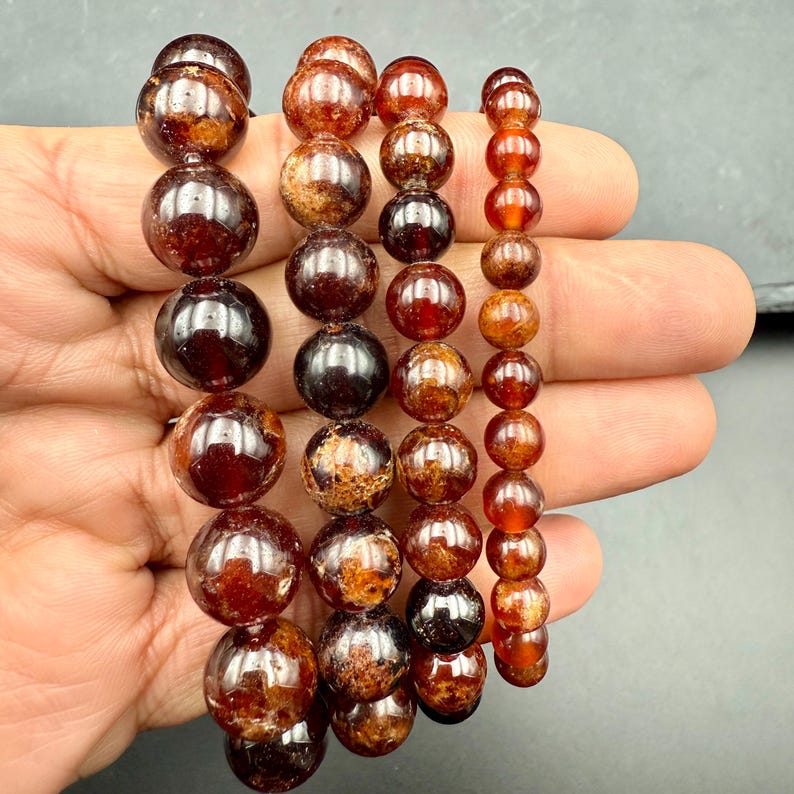 Pode incluir: Quatro pulseiras de contas com pedras de granada polidas, esf&eacute;ricas e de um tom castanho-avermelhado profundo. As pulseiras variam no tamanho das contas, desde esferas grandes, escuras e transl&uacute;cidas &agrave; esquerda at&eacute; contas mais pequenas e uniformes.