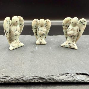 May include: Three small angel figurines carved from a speckled, light green stone. Each angel has outstretched wings and is standing on a dark gray surface. The angels are a light green color with black and brown speckles.