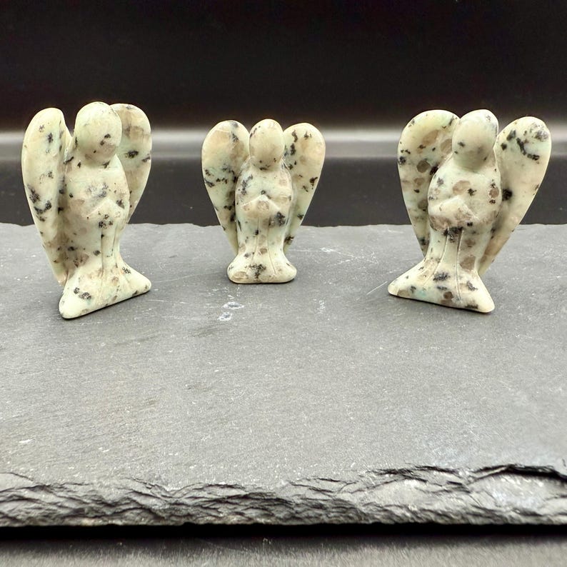 May include: Three small angel figurines carved from a speckled, light green stone. Each angel has outstretched wings and is standing on a dark gray surface. The angels are a light green color with black and brown speckles.