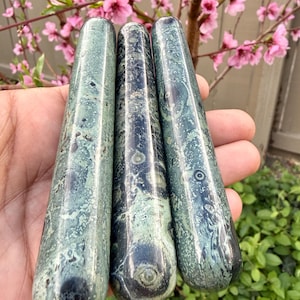 Puede incluir: Tres varitas de piedra verde y negra con puntas redondeadas. Las varitas tienen un aspecto veteado con remolinos verdes oscuros y negros. Las varitas se sostienen en una mano, con un fondo de flores rosas y follaje verde.