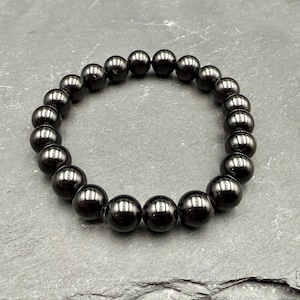 May include: A dark, glossy hematite bead bracelet. The bracelet features round, polished beads strung together, creating a continuous loop. The beads have a deep, reflective sheen, and the overall design is simple and elegant.