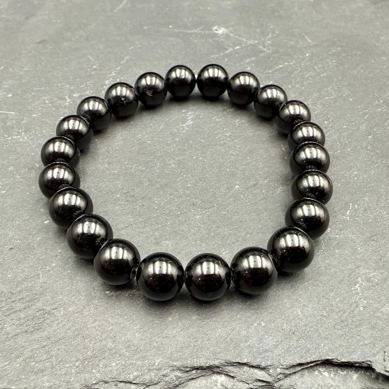 May include: A dark, glossy hematite bead bracelet. The bracelet features round, polished beads strung together, creating a continuous loop. The beads have a deep, reflective sheen, and the overall design is simple and elegant.