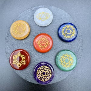 May include: Seven chakra stones in various colors, including red, orange, and blue, arranged in a circle on a gray surface. Each stone features a gold-colored chakra symbol. The stones are round and appear to be made of polished gemstones.