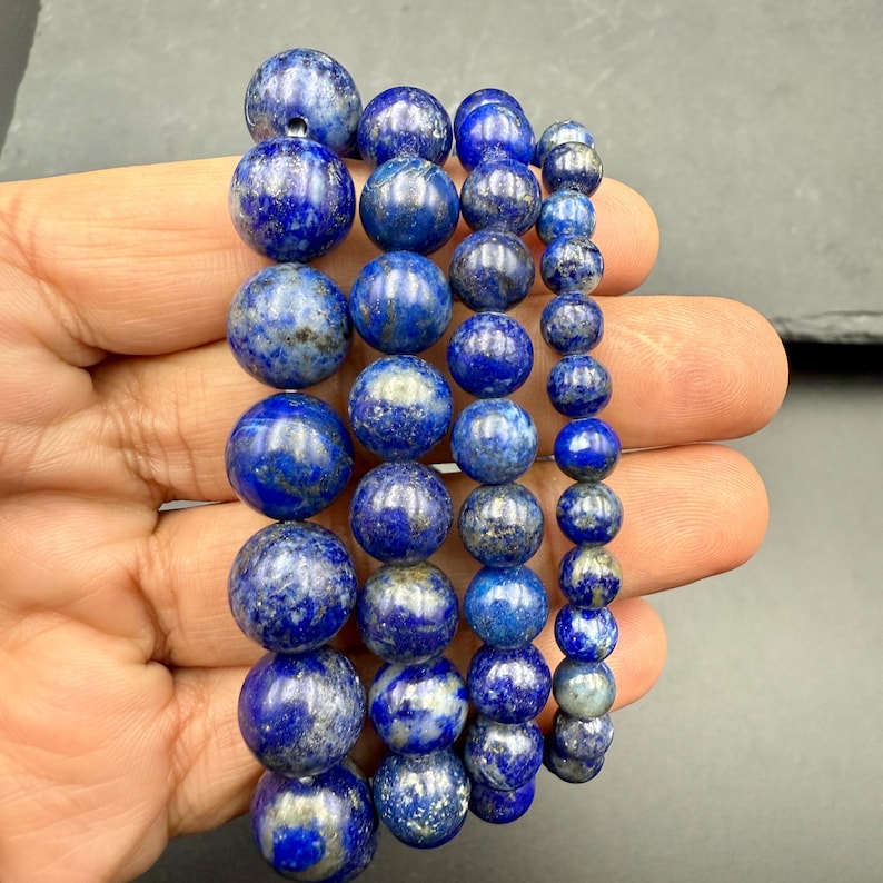 May include: A collection of four strands of round lapis lazuli beads arranged in descending order of size. The beads are deep blue with visible gold-colored pyrite inclusions and white mineral streaks, polished to a smooth, reflective finish.