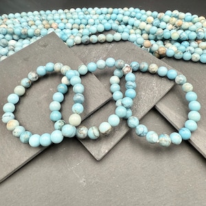Puede incluir: Tres pulseras circulares hechas de cuentas redondas de color azul claro con patrones de vetas naturales en marr&oacute;n y blanco. Las cuentas tienen un acabado liso y pulido y est&aacute;n ensartadas en un cord&oacute;n el&aacute;stico. Se observa una gran colecci&oacute;n de cuentas sueltas a juego.