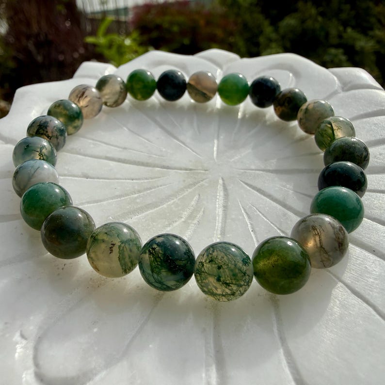 May include: A circular bracelet composed of polished, spherical moss agate beads. The beads feature a translucent to opaque appearance with varying shades of deep forest green, sage, and milky white, displaying intricate, fern-like dendritic inclusions.