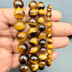 May include: Four beaded bracelets featuring tiger's eye gemstones. The bracelets vary in bead size, ranging from large to small. The beads display a mix of golden brown and dark brown colors, with a polished, reflective surface. The bracelets are strung together.