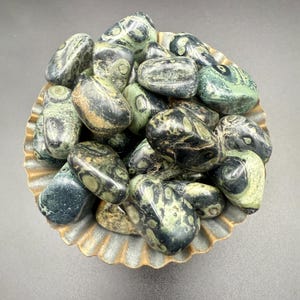 May include: A collection of tumbled stones in shades of green, black, and tan, filling a decorative, scalloped-edge bowl. The stones have unique patterns and textures, creating a natural and earthy aesthetic. The bowl has a metallic gold finish.