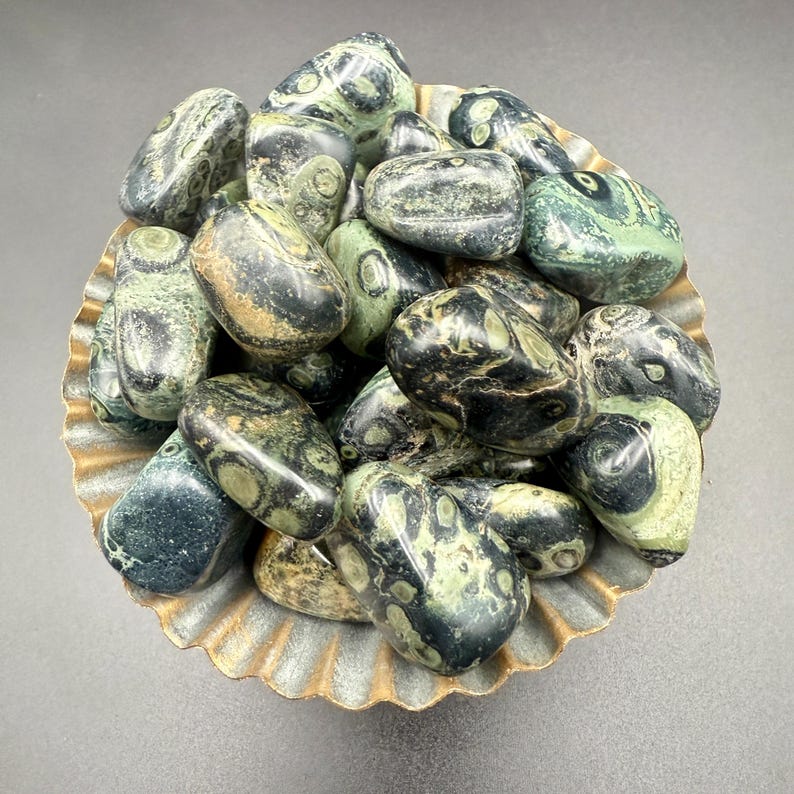 May include: A collection of tumbled stones in shades of green, black, and tan, filling a decorative, scalloped-edge bowl. The stones have unique patterns and textures, creating a natural and earthy aesthetic. The bowl has a metallic gold finish.