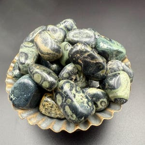 May include: A collection of polished leopard skin jasper stones in a decorative, scalloped-edge bowl. The stones are primarily green and black with unique spotted patterns. The bowl has a gold-colored rim and a textured surface.