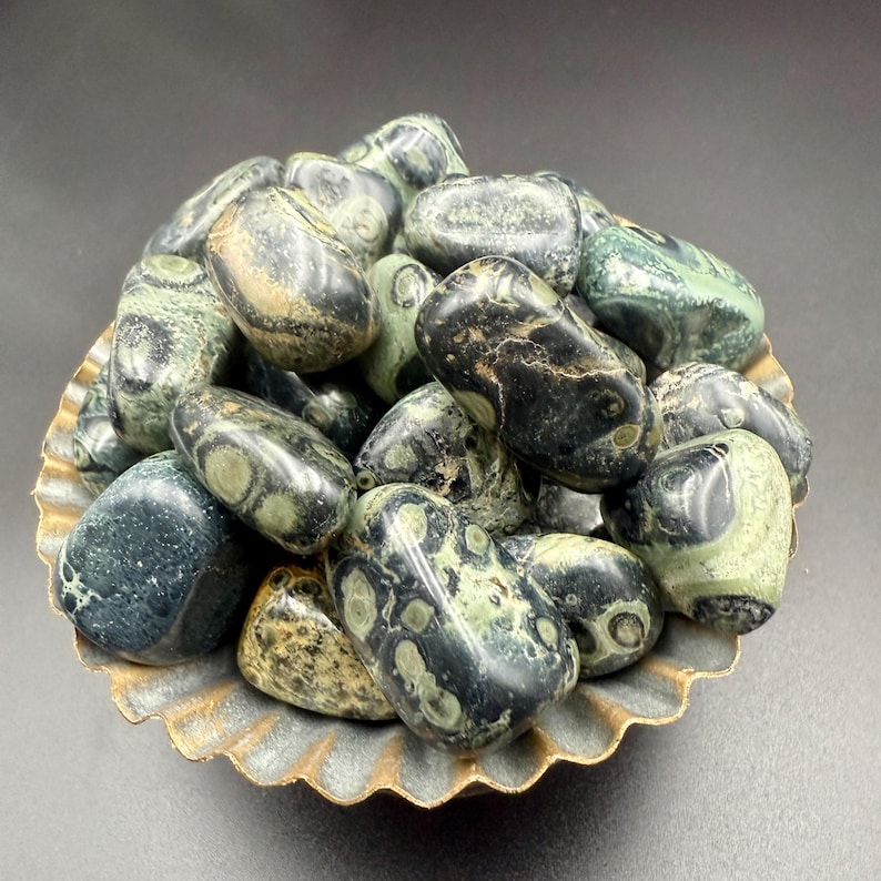 May include: A collection of polished leopard skin jasper stones in a decorative, scalloped-edge bowl. The stones are primarily green and black with unique spotted patterns. The bowl has a gold-colored rim and a textured surface.