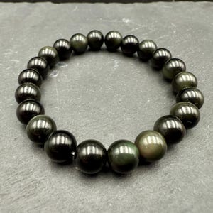 Puede incluir: Un brazalete de cuentas negras y verde oscuro. El brazalete está hecho de cuentas redondas pulidas con un brillo reflectante. Las cuentas están ensartadas para formar una forma circular. El brazalete se exhibe sobre una superficie gris.