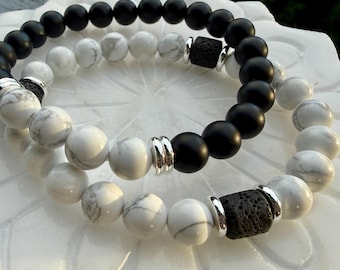 Puede incluir: Dos pulseras de cuentas. Una pulsera presenta cuentas negras, la otra cuentas blancas y grises veteadas. Ambas pulseras tienen detalles plateados y una cuenta de piedra de lava negra. Las pulseras están sobre una superficie blanca con forma floral.